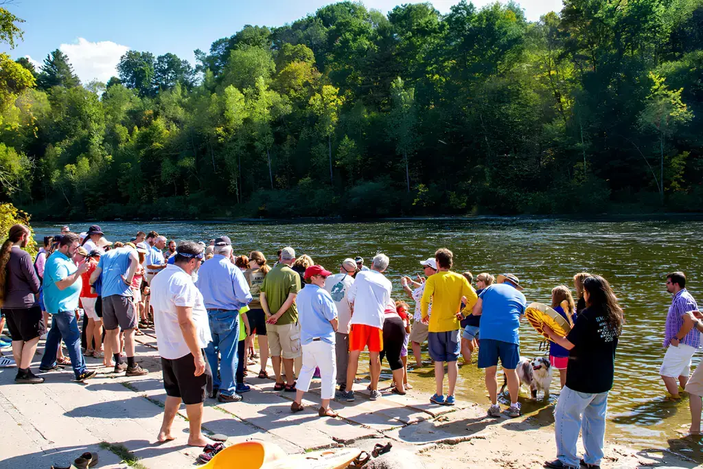A large group of people, some with dogs, gather on a riverside dock and sandy shore under a sunny sky, surrounded by lush green trees, watching something in the water.