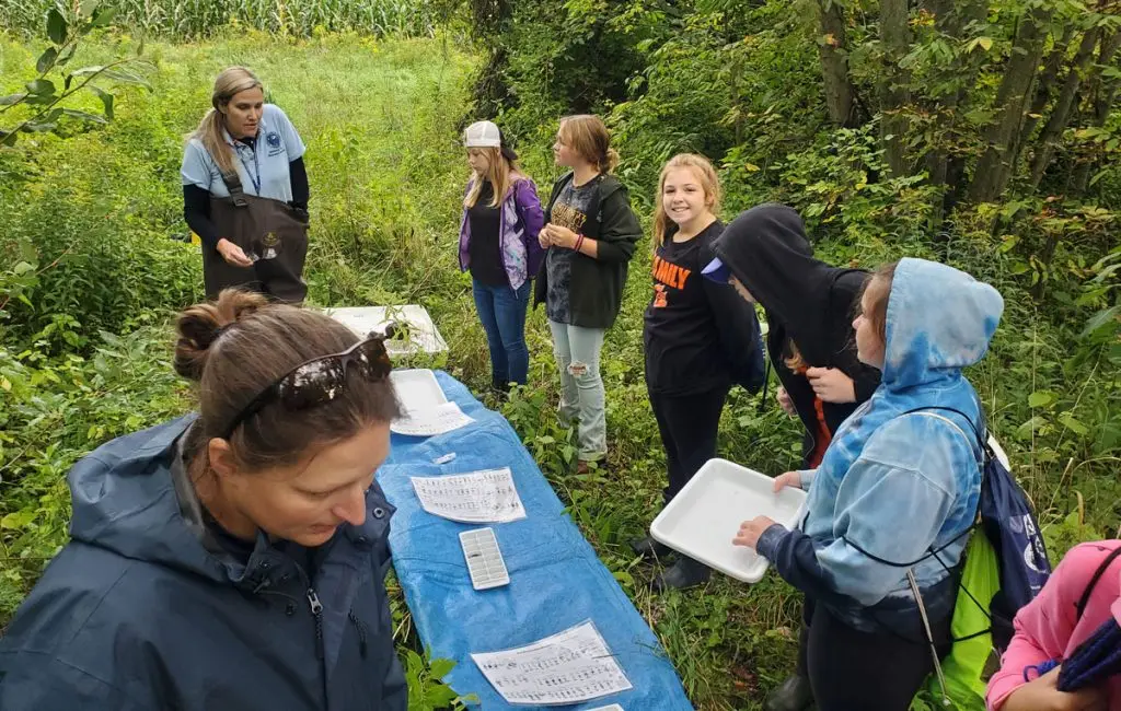 A group of people, mostly children, gather outdoors by a long blue table covered with papers and trays, engaged in a nature or science activity surrounded by dense greenery.