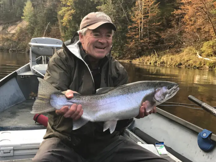 An older man in a rain jacket sits in a boat on a river, smiling and holding a large fish with both hands. Trees with autumn foliage line the riverbank in the background.
