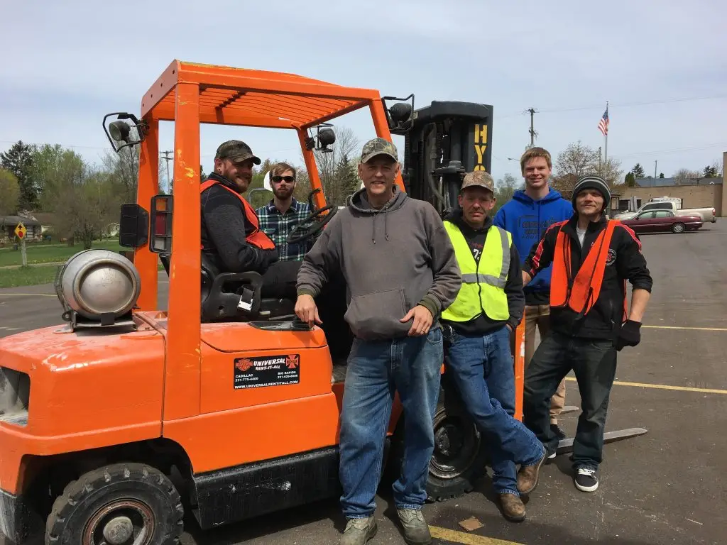 Six men wearing casual clothes and safety vests stand and sit around an orange forklift in a parking lot, smiling at the camera. Trees and buildings are visible in the background.