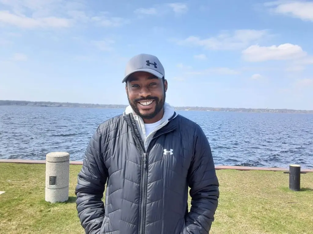 A man wearing a gray cap and a black Under Armour jacket stands smiling near a body of water with grassy ground and a cloudy sky in the background.