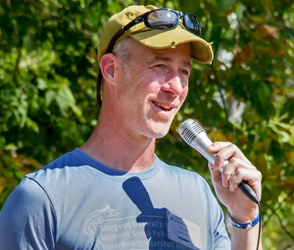 A man in a tan cap and sunglasses, wearing a blue shirt, holds a microphone and speaks outdoors. Green foliage is blurred in the background.