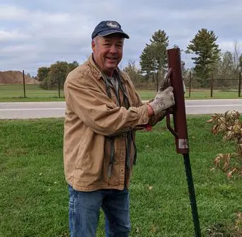 A man in a tan jacket and blue jeans smiles while using a post driver to install a metal fence post outdoors, with green grass, trees, and a road in the background.