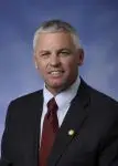 A man with short gray hair in a dark suit, white shirt, and maroon tie, smiling at the camera against a blue background.