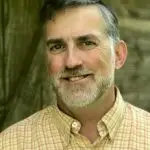 A middle-aged man with short gray hair and a trimmed beard, wearing a light yellow plaid button-up shirt, smiles while standing outdoors in front of a blurred, earthy background.