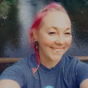 A person with pink hair, braided to the side, smiles while taking a selfie outdoors near a river. They are wearing a blue t-shirt and earrings. The background is blurred but shows water and greenery.
