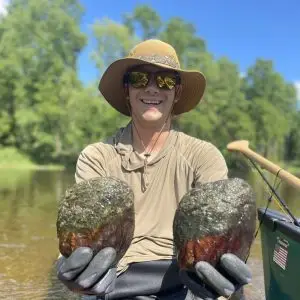 A person wearing a wide-brimmed hat, sunglasses, and gloves smiles while holding two large, round, mossy rocks by a river, surrounded by trees on a sunny day.