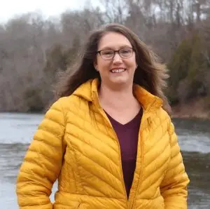 A woman with long brown hair and glasses, wearing a yellow jacket and a maroon top, stands smiling by a river with bare trees in the background.