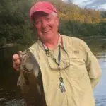 An older man wearing a red cap and beige shirt smiles while holding up a fish by a river, with trees and blue sky in the background.