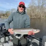 A person wearing a red cap, sunglasses, and a gray jacket stands on a boat holding a large fish, with a river and leafless trees visible in the background.
