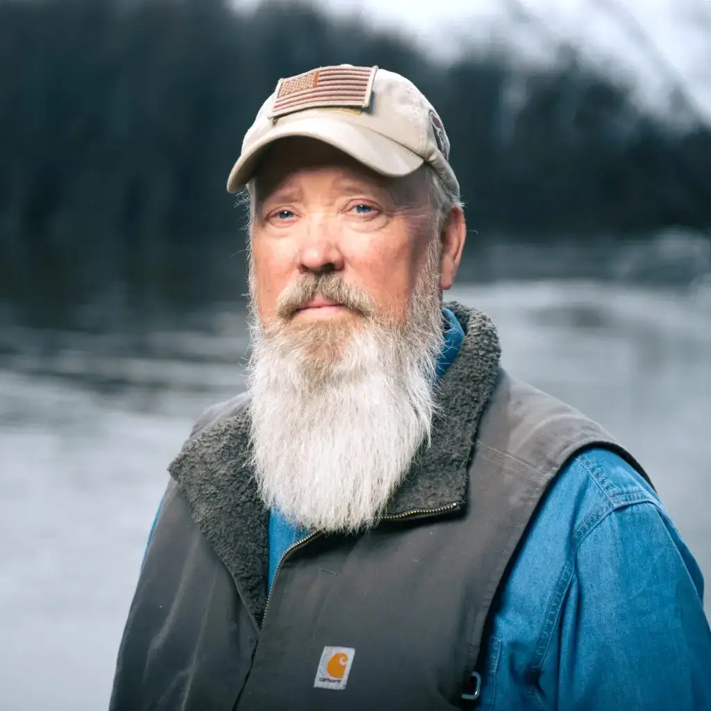 A bearded man wearing a tan cap with an American flag patch and a blue shirt stands outdoors near a body of water, with trees blurred in the background.