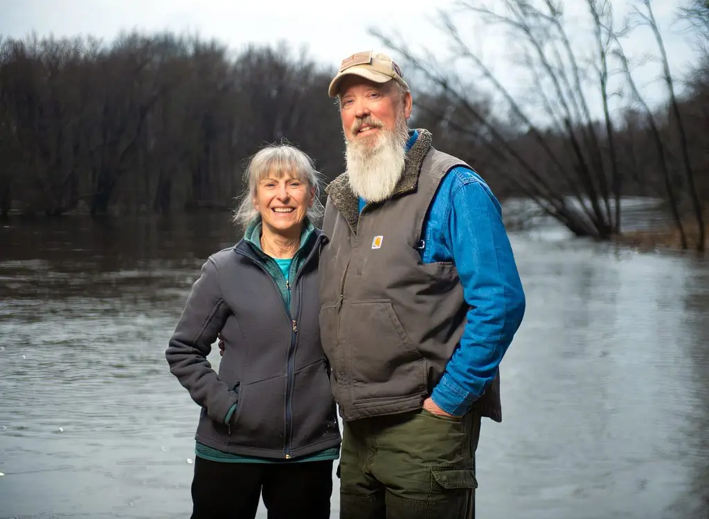 An older woman and man stand together outdoors by a river, smiling at the camera. Bare trees line the riverbank in the background, and both wear casual, outdoor clothing.